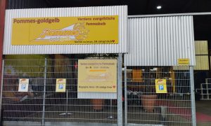 Eingangsbereich der evangelischen Pommesbude „Pommes Goldgelb“ in Verden mit gelben Schildern und Öffnungszeiten.