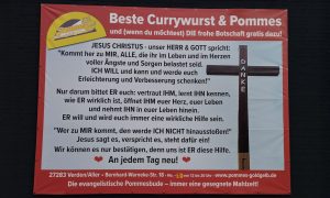 Banner der evangelischen Pommesbude „Pommes Goldgelb“ in Verden mit christlicher Botschaft und Aufschrift „Beste Currywurst & Pommes – und die frohe Botschaft gratis dazu“.